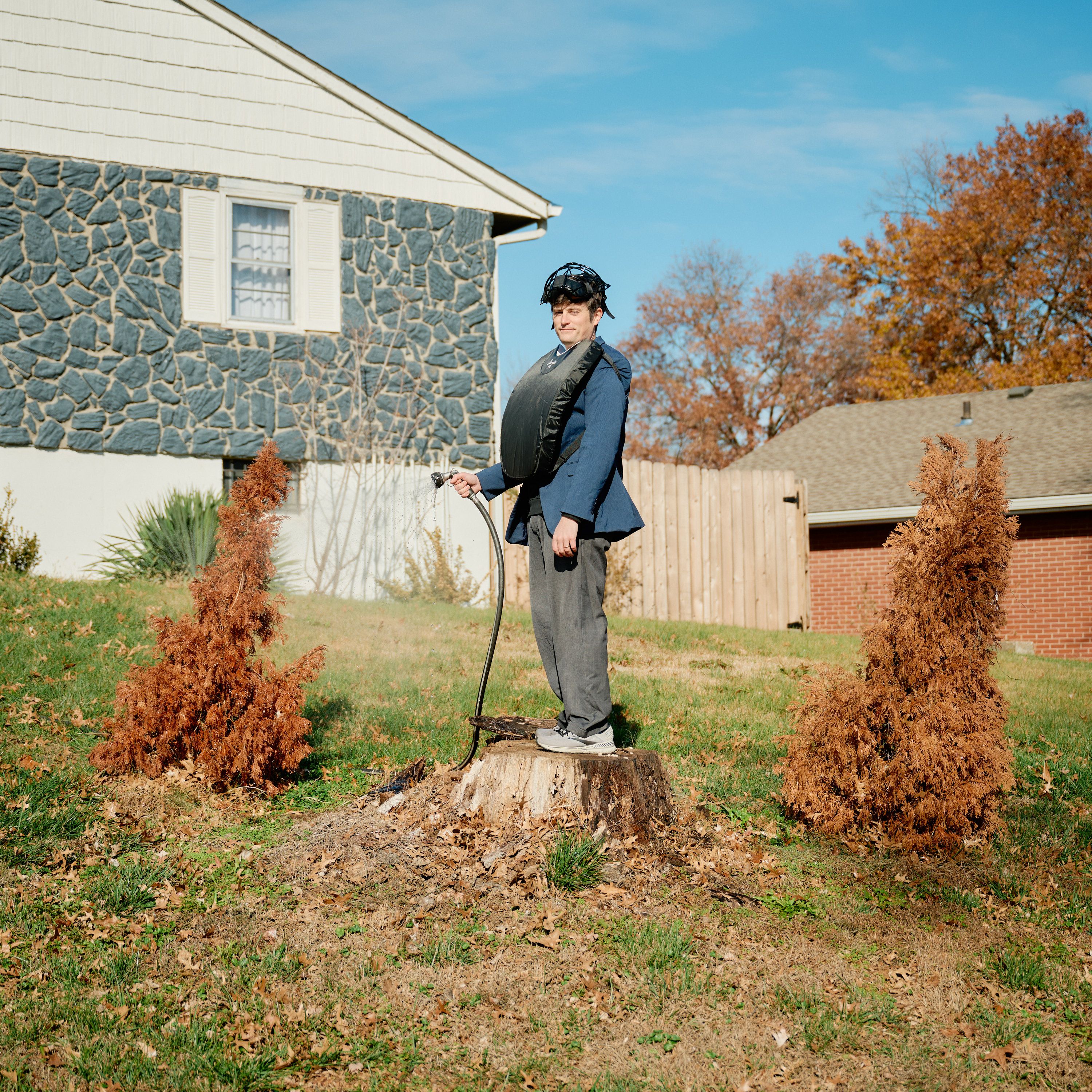 A man dressed as an umpire standing on a tree stump between two dead arborvitae trees, one of which he is watering with a garden hose. This photo was taken by David Robert Elliott.